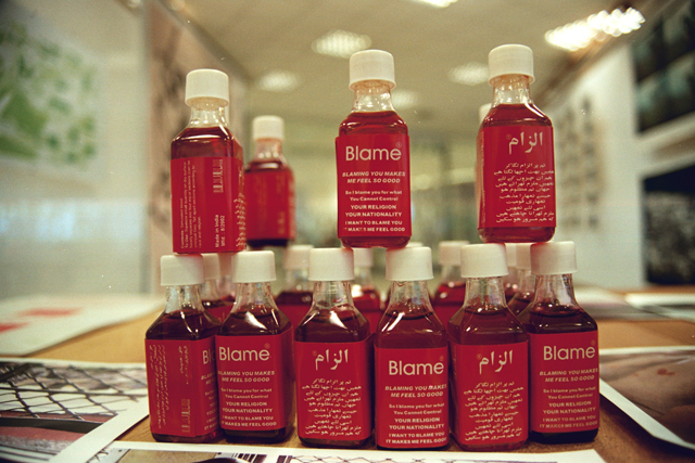 Shilpa Gupta. Blame, 2002–04. Interactive installation with Blame bottles that contain simulated blood, posters, stickers, video, interactive performance, 1 min 49 sec loop. Installation 300 x 130 x 340 cm (118 x 51 x 134 in).