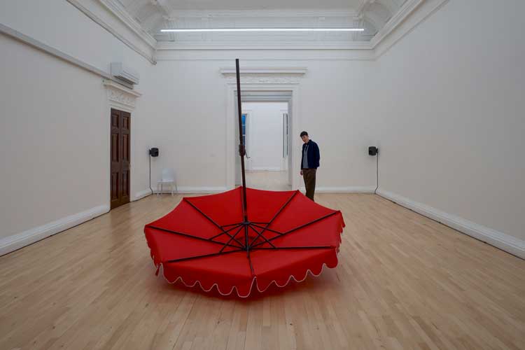 Laura Lima, Parasol Deux, 2023/2026. Red parasol, metal, motors, electrical components, wheels, sound. Audio composition by Laura Lima and Ricardo Siri. Installation view, Laura Lima: The Drawing Drawing, Institute of Contemporary Arts, London. Photo: Anne Tetzlaff.